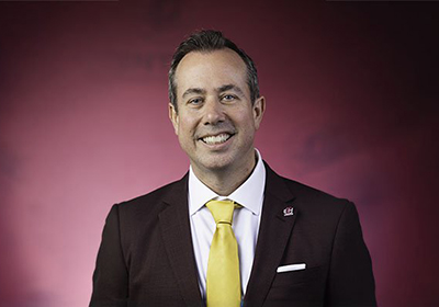CMU President Neil MacKinnon, PhD wearing a gold tie, stands in front of a maroon backdrop