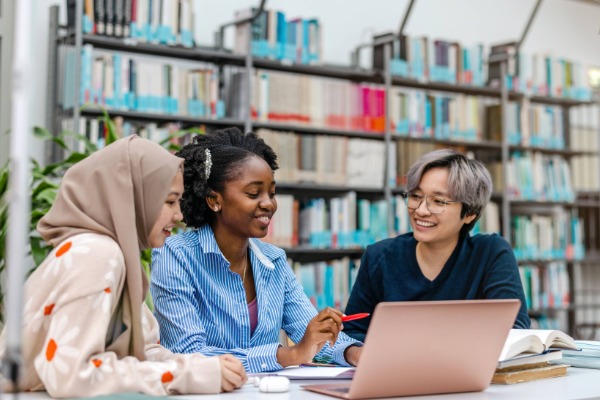 three students of different ethnicities sitting together looking at a computer. The background features stacks, or large shelving of books and resources.