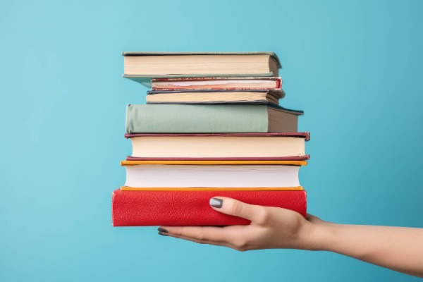 Photo has a medium blue solid background and the focus is a hand holding out a stack of books.