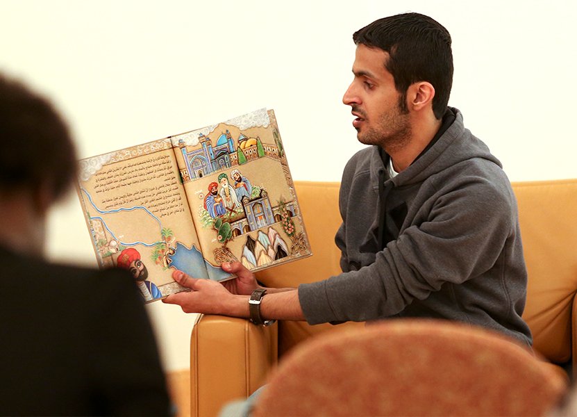 a man sitting in an orange leather chair holding a children's book with colorful pictures and reading to an audience
