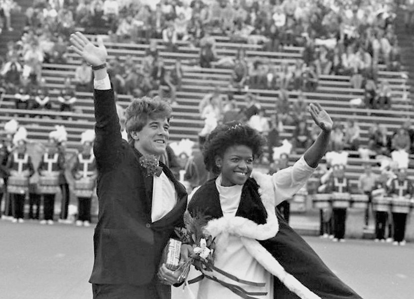 a man and woman waving with their arms up dressed as CMU Homecoming king and queen. A marching band and stadium seats full of people are in the background