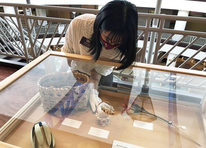 a woman wearing a mask and gloves placing woven baskets to display in an exhibit case