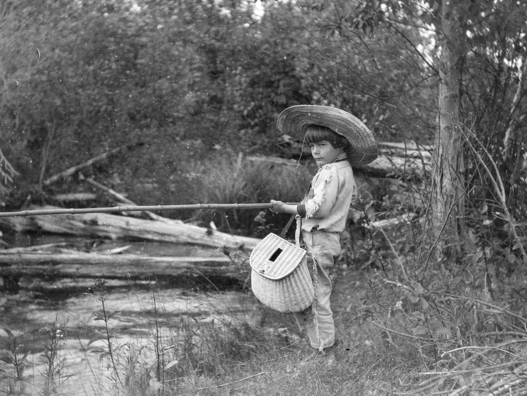 Hemingway as a young boy fishing next to a creek, dressed in large straw hat, a long sleeve shirt, long pants and holding a woven straw bag