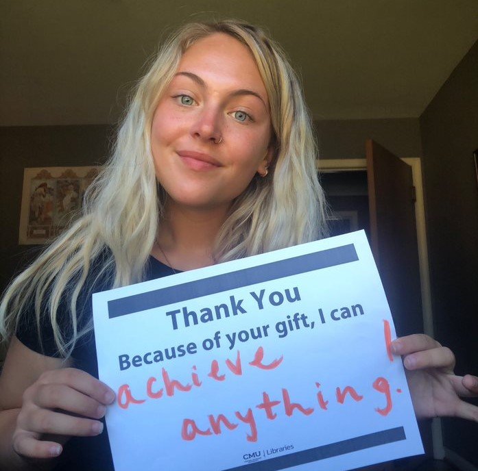 Student Scholarship recipient smiling and holding a blue and white poster that says, Thank you, because of your gift, in blue font and, I can achieve anything, written in orange marker