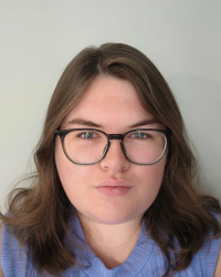 A woman with brown hair and black glasses wears a blue blouse and smiles in front of a grey backdrop in selfie type headshot.