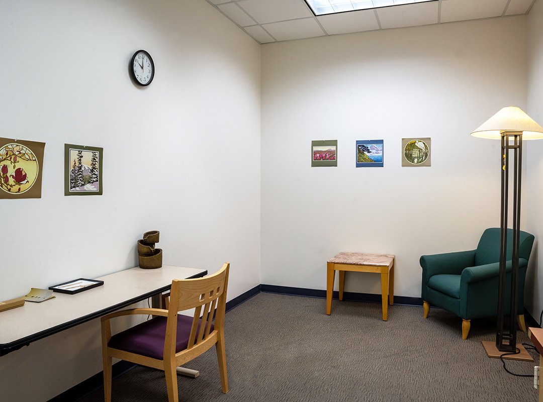 Image of two walls in the Quality of Life Room at the CMU Park Library. Walls are white with a few pictures, furniture featured, slim table, wood chair with arm, end table, lamp, and cushioned green chair