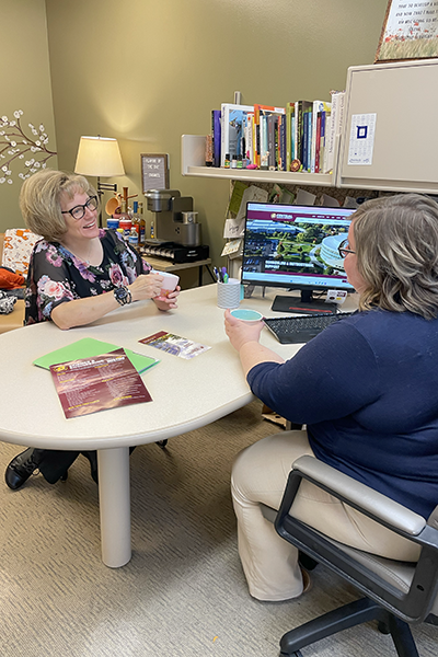 Two individuals seated across from one another at a desk in an office and talk to each other