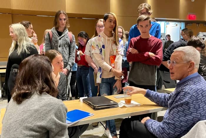 a group of people students standing around a table listening to a mentor talk to other students