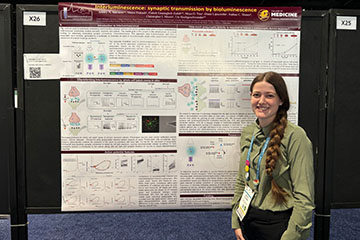 A woman with a long brown braid wearing a green blouse stands in front of a large research poster.
