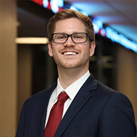 Trevor_Carnovsky_200x200 Trevor Carnovsky smiling in a professional headshot, wearing glasses and a suit with a red tie in an indoor setting.
