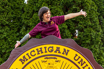 Alisha Draper sits on top of the CMU seal in Warriner Mall. Her hands are outstretched and she has an exaggerated grin. She wears a maroon CMU polo shirt. She has short brown hair.