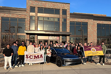 Central Michigan University students pose with a Lucid Air electric vehicle outside the College of Business Administration building during a Supply Chain Management Association event.