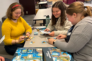 Three young women gather around a table and review the game components for Hydrologic Cycle. They are holding cards and an instruction sheet. The game board sits in front of them.