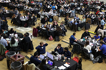 Students and industry professionals sit at round tables during Central Michigan University’s ERPsim competition, collaborating in teams while using laptops and business simulation software.