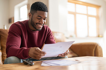 A student in a maroon long sleeve shirt and jeans sits at a coffee table and reviews financial documents. He is looking at them and smiling. He has a beard and short hair.