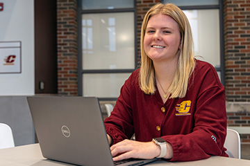 Hannah Everitt sits at a table in the Grawn Atrium. She's wearing a long-sleeve maroon cardigan with an Action C logo. She has long blond hair and is smiling. In front of her is a laptop.