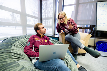 A student sits in an oversized green beanbag chair in the Idea Den. He has a long sleeve maroon CMU shirt on and has a laptop on his lap. In a chair next to him, a second student leans over and gestures with her hand. She wears a maroon CMU sweatshirt.
