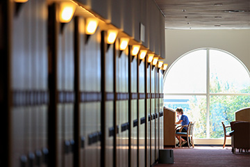 A student sits at a desks in front of an upper story window at Park Library. Book cases run alongside of the image on the left in the foreground.