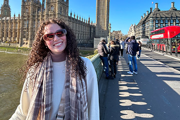 Rachel Meininger has long curly hair and wears a sweatshirt and scarf as she stands on a bridge in London. A red double decker bus is in the background.