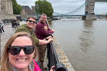 Faculty members Shelly Bartosek wearing sunglasses and a hot pink jacket and Kyle Nothstine wearing a CMU sweatshirt stand with a student in a green sweatshirt near the edge of the Thames river with a bridge in the background.