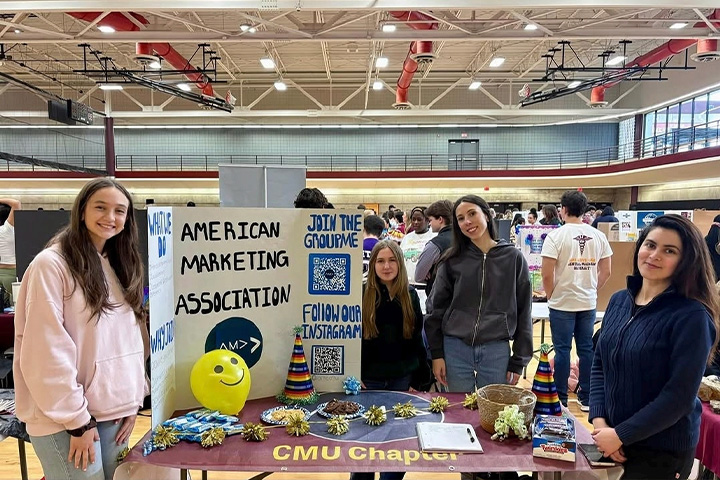 Four CMU students stand behind an American Marketing Association table during a campus organization event. Sarah Firchau stands on the far left, and Aimen Khan stands on the far right.