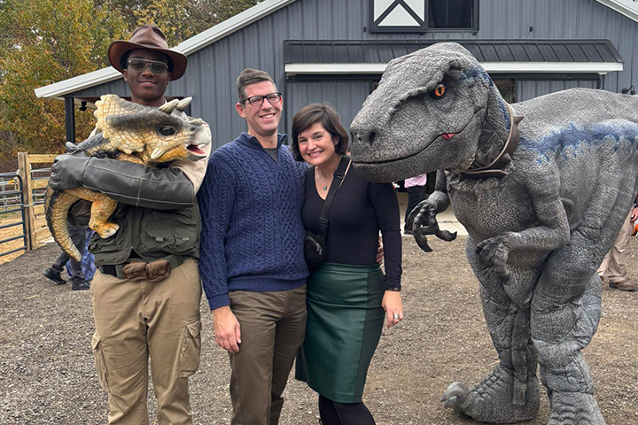 Alisha Draper stands with her partner and smiles. To her right is a large velociraptor and to her left is a man holding a baby triceratops. Alisha and her partner are dressed up, she's wearing a green skirt and black long-sleeve top.