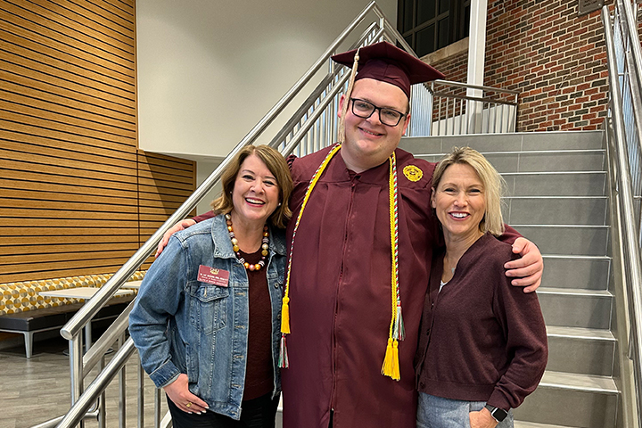 Amy McGuinnis (left), Blake Stanbaugh (center), and Lori Driessnack (right) stand in front of the Atrium stairs. Blake wears his graduation cap and gown and has his arms around Amy and Lori.