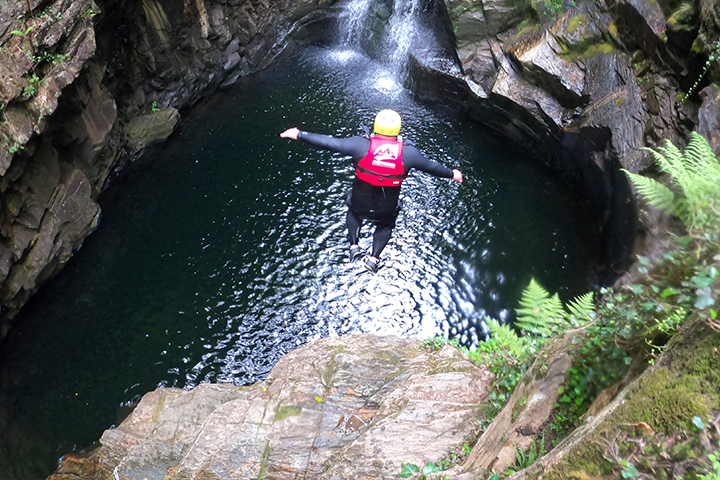 Blake Stanbaugh wears a wetsuit, life jacket and helmet as he jumps off a rocky outcropping into water far below.