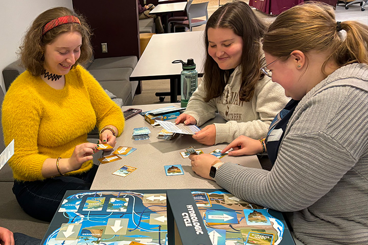 Three young women gather around a table and review the game components for Hydrologic Cycle. They are holding cards and an instruction sheet. The game board sits in front of them.