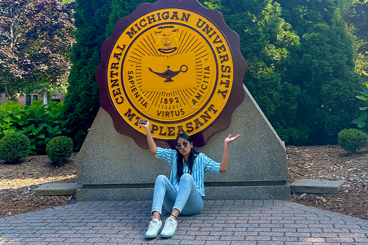Maheshwari Kammadanam sits on brick pavers outside in front of the CMU seal on Warriner Mall. Behind the sear are evergreen bushes. She's wearing a blue and white striped shirt, sunglasses, jeans and white tennis shoes with an ankle bracelet.