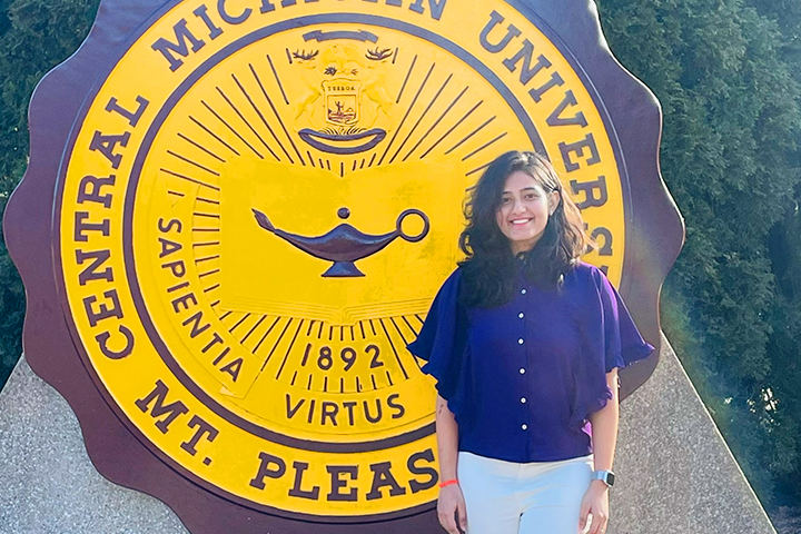 Pravalya Kamireddy stands in front of the large maroon and gold seal in front of Warriner Hall. She's wearing a blue button down shirt with loose angled sleeves, white pants, and an Apple watch. Her long dark hair is pulled behind one ear and she smiles.