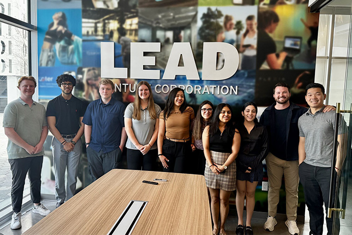 A group of students in the Wakeling-Gendron Entrepreneurial Scholars program pose with LEAD Venture Corporation representatives in a boardroom during a visit. In front of them is a large table and behind them is a wall graphic that reads