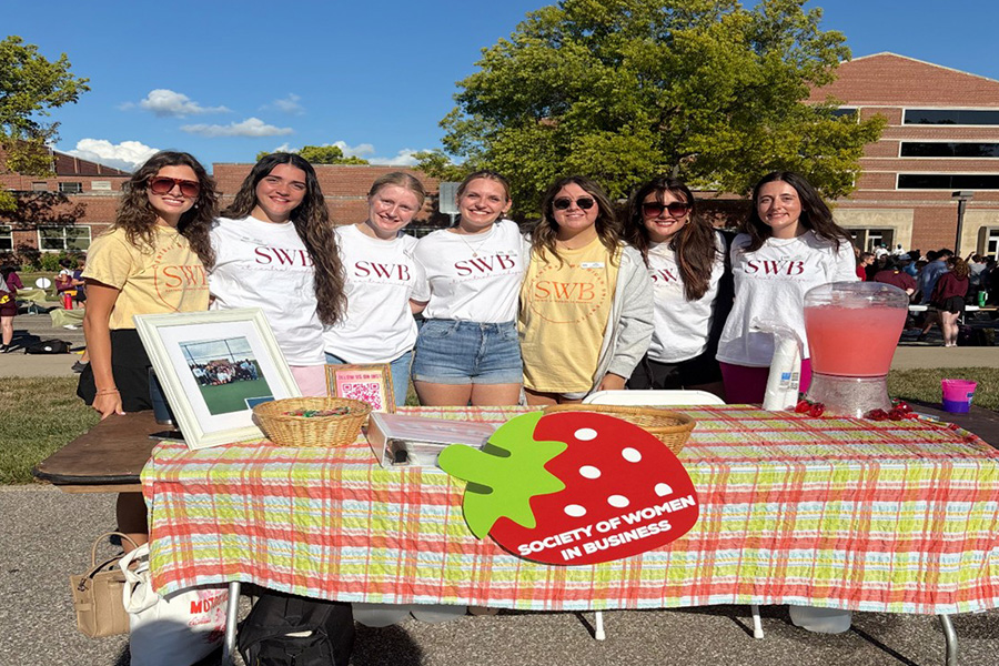 Members of the Society of Women in Business stand behind their table at the fall Mainstage event. They are all wearing SWB shirts and have their arms around each other. Carmella Cortez stands as the second to the right member of the group of seven women.