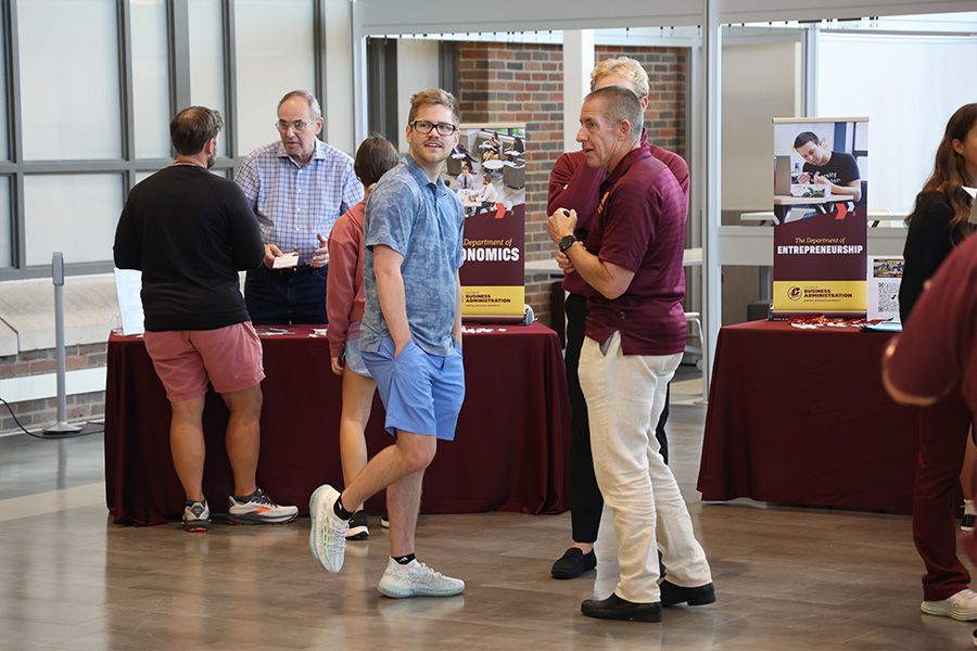 900x600_Article_Inset_CMUandYou_1 A prospective student wearing blue shorts and a blue polo shirt talks with a faculty member in a maroon CMU polo shirt and khaki pants in the Grawn Atrium. Behind them are tables with banners for the CBA departments.