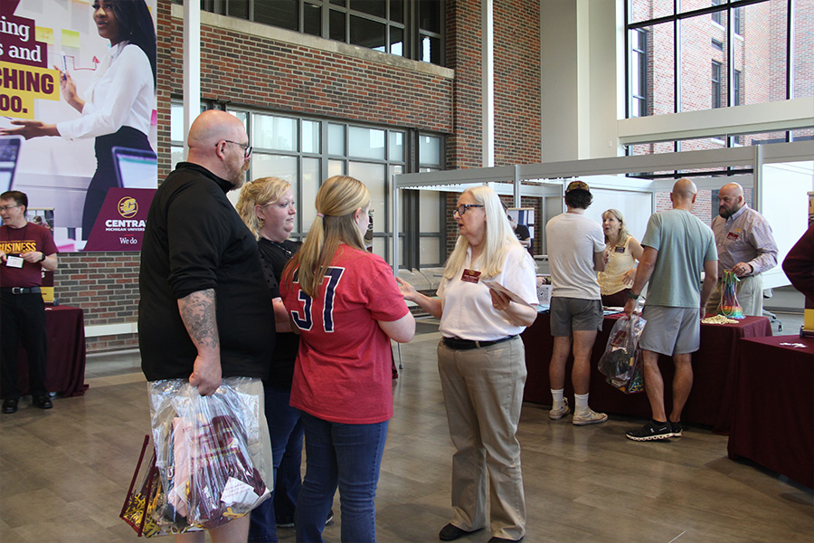 900x600_Article_Inset_CMUandYou_2 A staff member in a white polo shirt and khaki pants holds brochures and speaks with a family in Grawn Hall. In the background are families visiting information tables for each department.