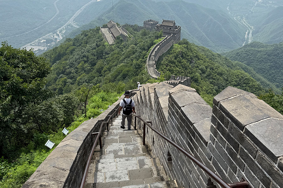 A single man with a white tshirt, gray pants, hat and black backpack walks down the steps on the Great Wall of China. The wall winds into the distance, surrounded by hills and forest. 