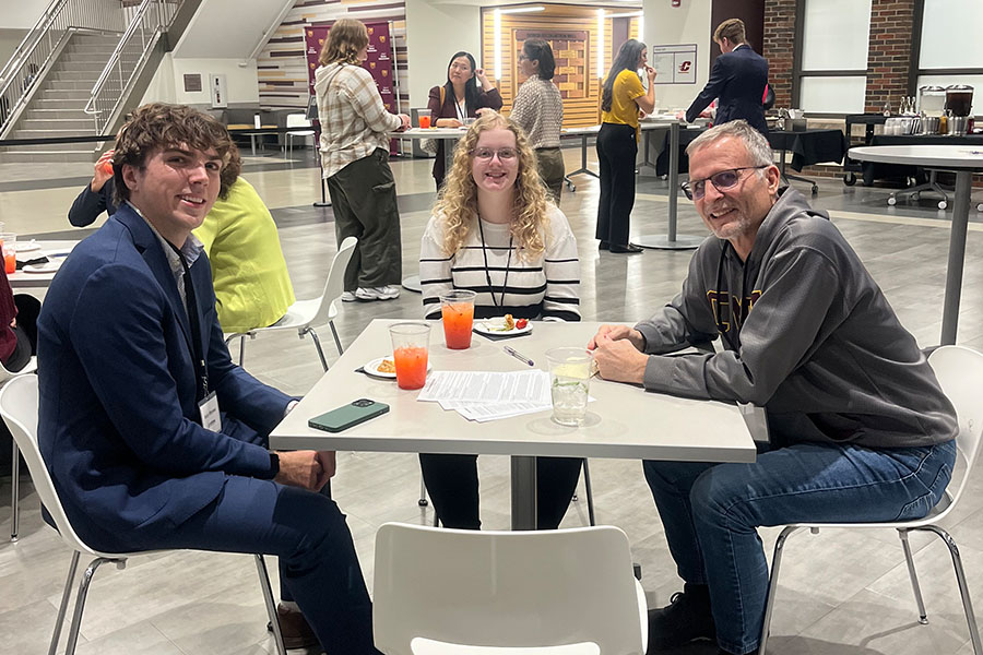 Left: a male student with wavy hair wears a navy suit. Center: a female student with long blonde wavy hair wears a white and black striped shirt with black pants. Right: a faculty member wears a CMU sweatshirt and jeans. The are seated with mocktails.