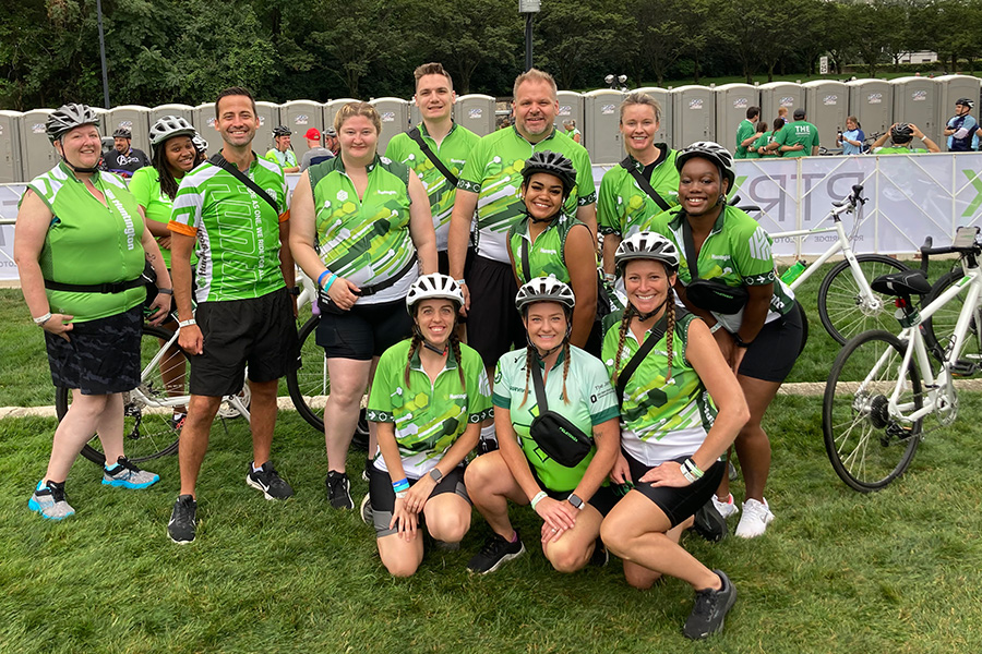 Jason Blik stands with eleven other team members on the Huntington Bank bike team. They are all wearing green Huntington bank cycling shirts and black pants. Some still wear helmets and many have black crossbody bags. Their bikes stand to the side.