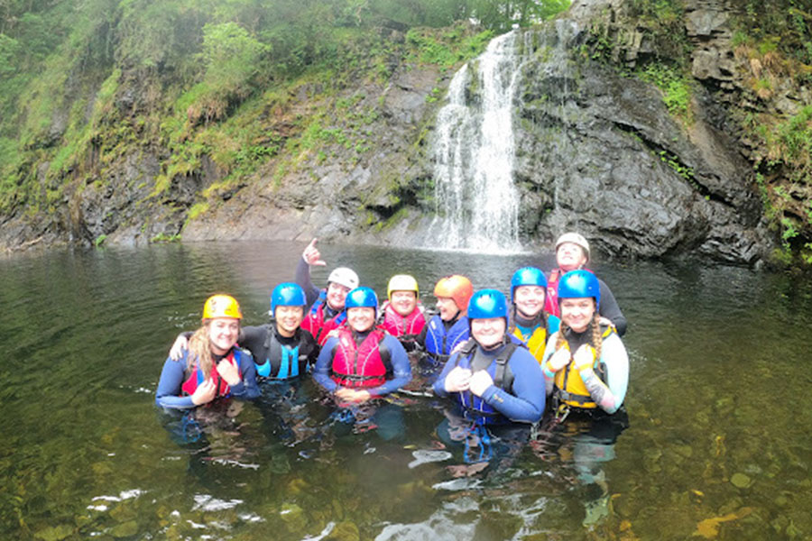 A group of CMU students stand in a river wearing colorful life jackets and helmets in front of a waterfall.