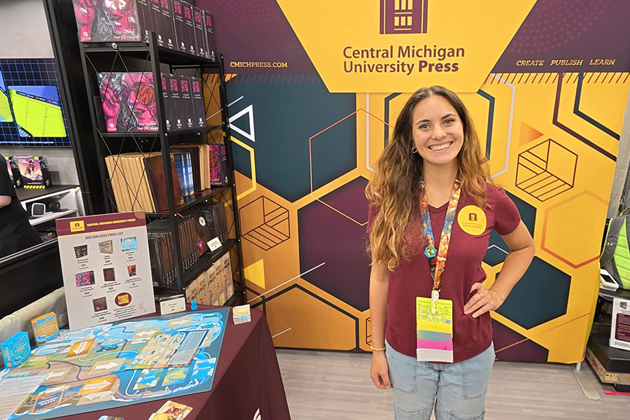 Lauren Bonomo stands in the CMICH Press game booth at Gen Con. She's wearing a maroon CMICH Press t-shirt with a convention pass on a colorful lanyard. She is surrounded by tables and shelves with games published by the press.