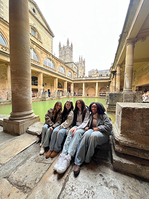 Rachel Meininger sits with three other students at an historic roman bathhouse with a pool and marble pillars in the UK.