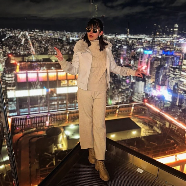 Aimen Khan stands on a platform overlooking a brightly-lit cityscape. She's wearing a tan sweater, tan ankle length pants, light brown boots with a chunky heel, and a white cream jacket with a fur color. She has black sunglasses. 