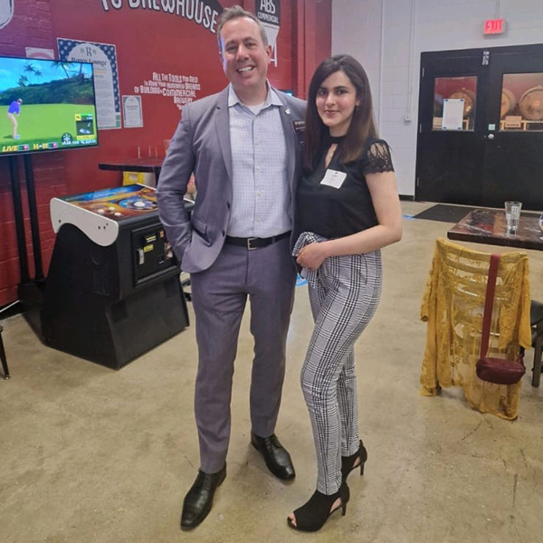 Aimen Khan stands with CMU President Neil MacKinnon. She wears a black short-sleeve shirt, black plaid pants and black heels. Pres. MacKinnon wears a gray suit. They are in a brewery with arcade games and casks in the background.