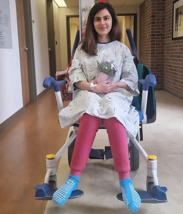 Aimen Khan sits in a wheelchair in a hospital corridor. She has long dark brown hair and smiles slightly. She wears a hospital gown over pink pants and wears blue hospital grippy socks. She holds a plant in her lap. 