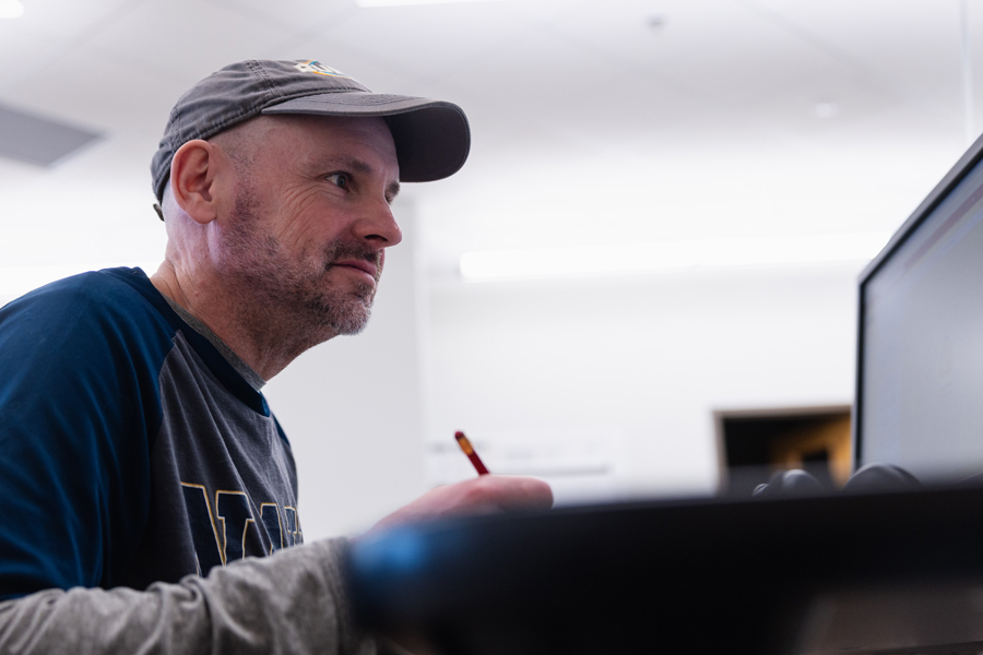 A man wearing a gray cap and navy long-sleeve shirt sits at a desk, holding a pencil and looking at a computer screen with a focused expression.