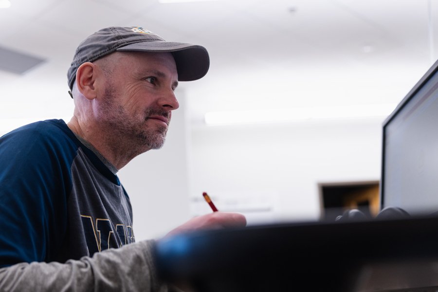 A man wearing a gray cap and navy long-sleeve shirt sits at a desk, holding a pencil and looking at a computer screen with a focused expression.