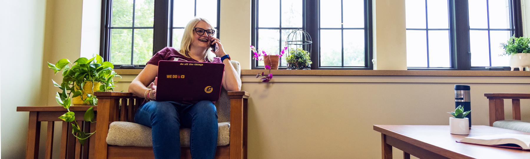 A smiling woman sits in a wooden chair by tall windows, talking on the phone with a maroon laptop on her lap. The laptop has stickers that say “WE DO” and “do all the things.” Plants and an open book decorate the cozy room.