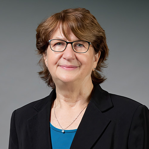 Jan Hewlett wears a blue shirt, black blazer, necklace and black glasses.
