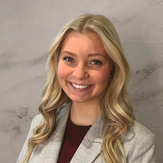 Camryn Asper wears a gray blazer over a maroon shirt. She has long blond hair and is smiling at the camera. Behind her is a photo backdrop that looks like gray marble.