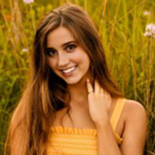 Caprice Radman poses in a field. She has on a light orange tank top. She has long brown hair and is smiling at the camera. She has one hand on her neck.